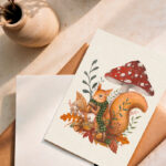 Carte illustrée “Un automne cosy” avec un écureuil tenant un gland, entouré de feuilles et d’un champignon rouge. Illustration automnale douce et poétique signée L’atelier de l’Écureuil.