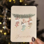 Carte illustrée représentant trois chaussettes de Noël suspendues à une branche de sapin décorée.
