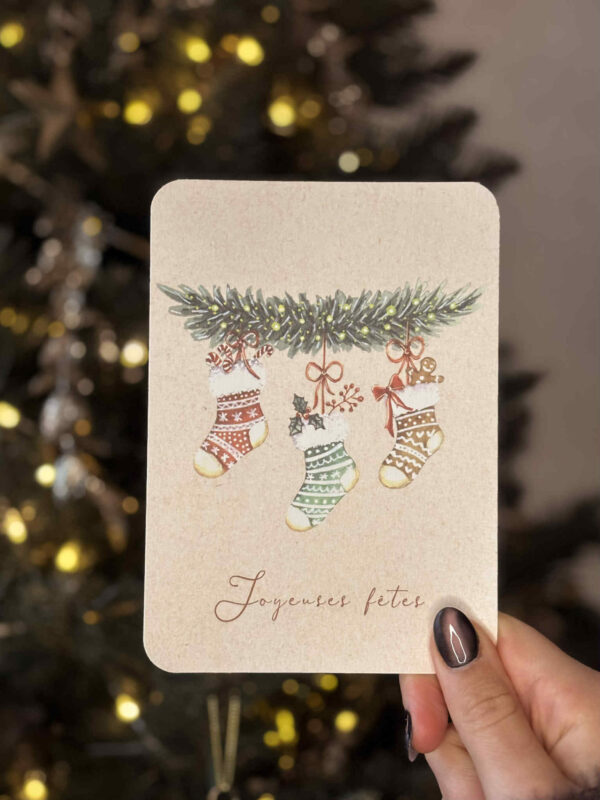 Carte illustrée représentant trois chaussettes de Noël suspendues à une branche de sapin décorée.