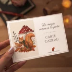Carte cadeau illustrée à l’aquarelle d’un écureuil roux sous une amanite, tenue en main devant un bureau éclairé à la bougie.